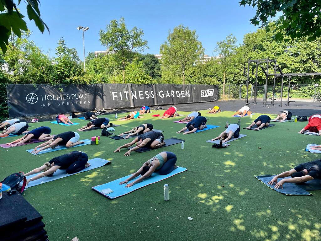 Yoga Teilnehmer praktizieren Vinyasa Yoga im Freien unter blauem Himmel und Sonnenschein in Düsseldorf bei Holmes Place am Seestern